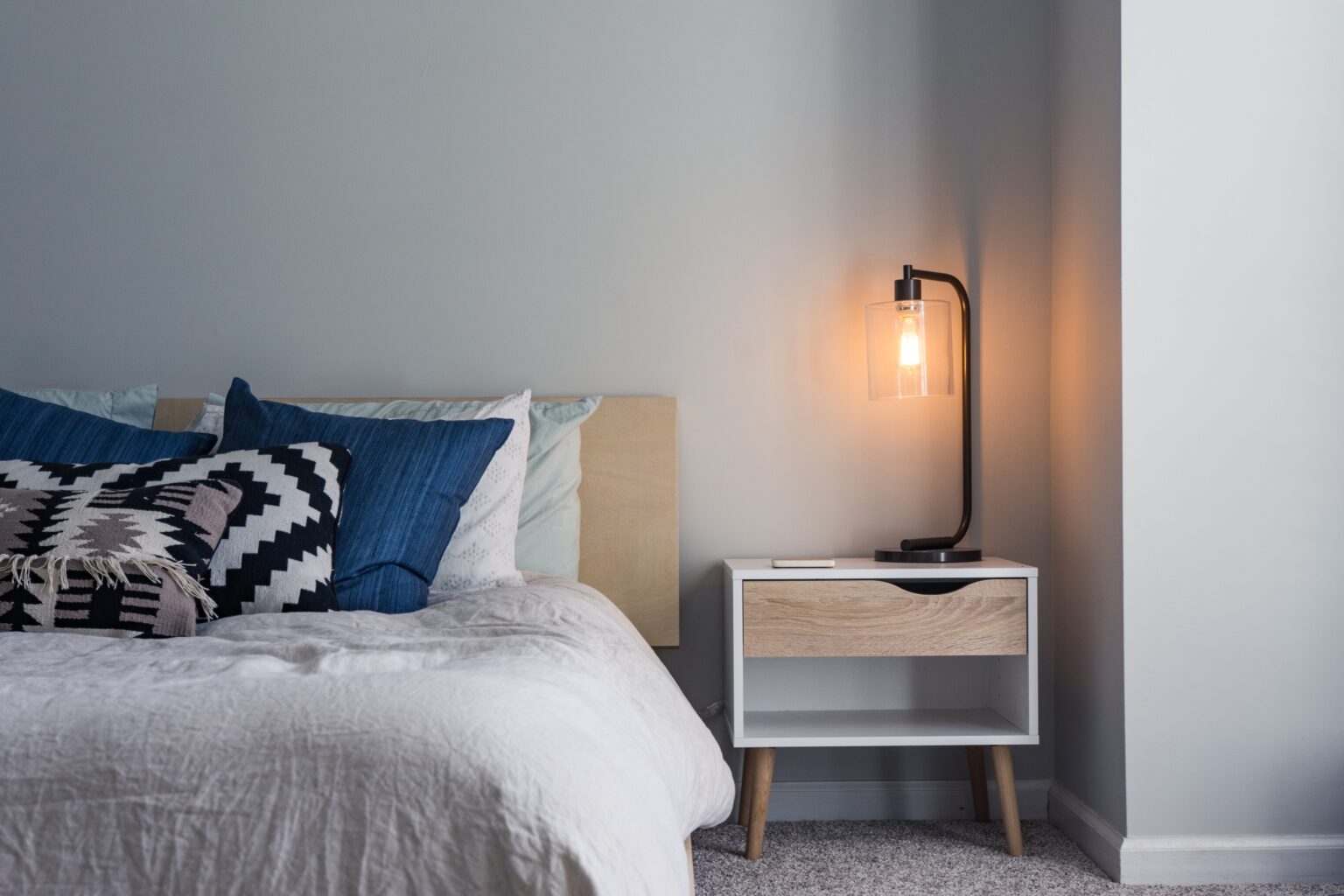 Photo of a bed and a nightstand with a light turned on.