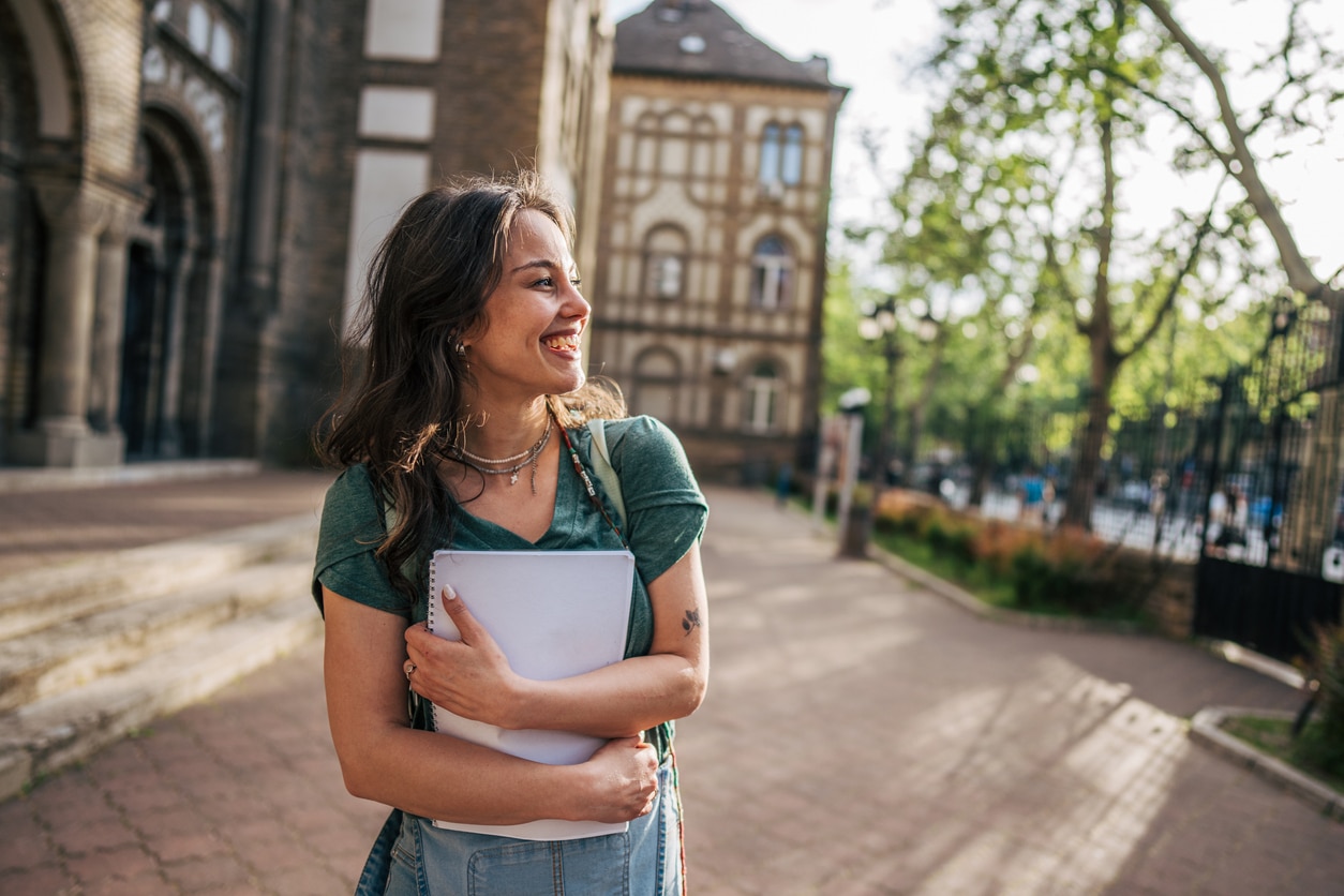Smiling university student walking outside college campus holding notebook.